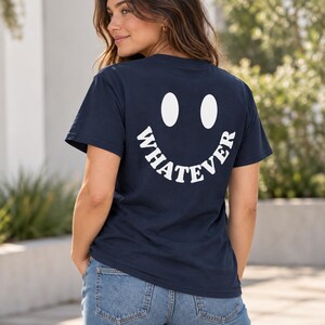 Pode incluir: Uma t-shirt de manga curta azul-marinho escuro com um grande gráfico branco nas costas. O design consiste em dois olhos ovais acima da palavra WHATEVER, impressa numa fonte branca curva e em negrito para formar um rosto sorridente.