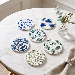Puede incluir: Un juego de cinco posavasos redondos de cerámica con patrones distintos: flores azules, corazones azules, hojas verdes con puntos naranjas, follaje azul, frondas de helecho verdes con bayas rojas y ramas frondosas sobre fondo blanco.