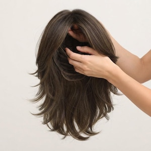May include: A medium-length brown wig with layered, textured ends and subtle lighter highlights throughout. The wig features a dark brown base color with multi-tonal strands, showcasing a visible interior cap construction with adjustable straps.