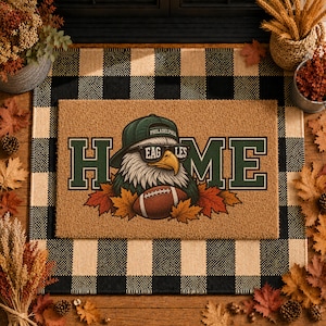 May include: A rectangular tan coir doormat featuring the word HOME in dark green block letters. The letter O is replaced by an illustration of a bald eagle wearing a green baseball cap and sunglasses, holding a brown football amidst autumn leaves.