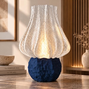 Può includere: Una lampada da tavolo con paralume in vetro trasparente ondulato dal design plissettato verticale. La base è un oggetto sferico blu scuro con una superficie testurizzata e sfaccettata. La lampada emette una luce calda dal paralume.