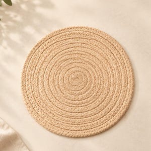 Könnte beinhalten: Ein rundes, beiges Platzset aus geflochtenem Seil mit einem spiralförmigen Design und sichtbaren Nähten. Der Text ELEGANT THREADS HEART Threads of Elegance, Made for You. steht in der oberen rechten Ecke über dem Platzset.