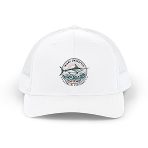 Op de afbeelding: Een witte truckerpet met een cirkelvormige afbeelding op de voorzijde. Het ontwerp toont een zwaardvis die over oceaangolven springt met een zonsopgang op de achtergrond, een zeester en de tekst MIAMI SWORDFISH COASTAL EXPLORER en EST. 1979.