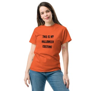 Op de afbeelding: Een feloranje T-shirt met korte mouwen en de zwarte tekst THIS IS MY HALLOWEEN COSTUME in een druipend, griezelig lettertype op de borst. Het kledingstuk heeft een ronde hals en een standaard pasvorm, gecombineerd met een blauwe spijkerbroek.