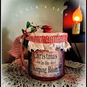 May include: A cylindrical container with a red lid, wrapped in a frayed beige fabric and a red and white striped ribbon. The label features the text "Christmas in the Keeping Room" in black font, accented by a small sprig of green leaves and red berries.