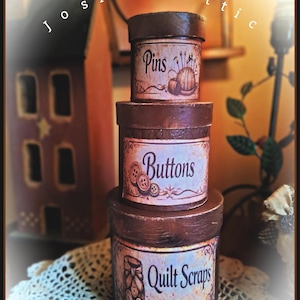 May include: A stack of three cylindrical brown storage boxes with lids. Each box features a label with decorative text and illustrations: the top box is labeled Pins, the middle box is labeled Buttons, and the bottom box is labeled Quilt Scraps.