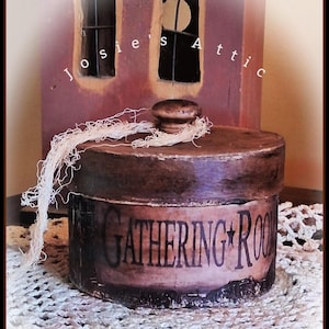 May include: A round wooden box with a dark, distressed finish and a matching lid featuring a small knob. The side of the box displays the text GATHERING ROOM in black lettering. A piece of frayed, light-colored string is tied around the lid knob.