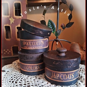 May include: A collection of antique, round, black wooden boxes with labels. The labels read "Spool Cotton," "Colors," and "J&P Coats." The boxes are stacked on a white, crocheted doily. The image also includes a decorative metal stand.