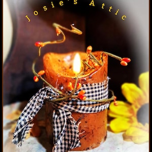 May include: A cylindrical orange candle with a lit wick, wrapped in a black and white checkered fabric ribbon and decorated with thin brown stems featuring small orange berries. The text Josie's Attic is printed in yellow above the candle.