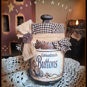 May include: A glass jar with a black and white checkered fabric lid cover, a tan burlap ribbon, and a decorative button. The label on the front features the text Grandma's Buttons with an illustration of buttons. The lid has a black knob handle.