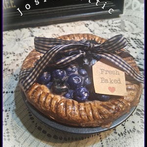 May include: A decorative pie with a brown crust, filled with blue berries, and tied with a navy and white checkered ribbon. A small tag reads "Fresh Baked" with a red heart. The pie sits on a dark gray plate.