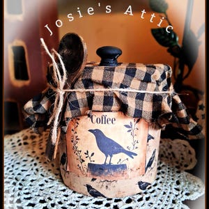May include: A cylindrical coffee canister featuring a tan label with a black crow illustration and the text "Coffee". The lid is covered in a black and tan buffalo check fabric, tied with twine and a small wooden spoon attached to the side.