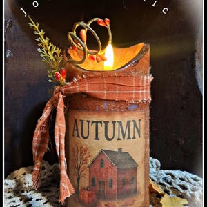 May include: A cylindrical candle with a textured brown finish, featuring the text AUTUMN and an illustration of a red house and pumpkin. It is wrapped with a frayed orange plaid fabric ribbon, a sprig of greenery, and red berries, with a lit flame.