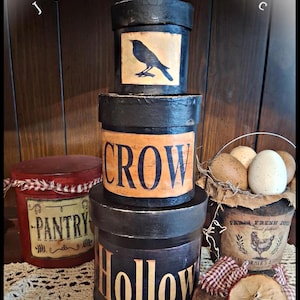 May include: A stack of decorative, round, black boxes with lids. The boxes are labeled "Crow" and "Hollow" in gold lettering. A red box with "Pantry" is also visible. A bucket of eggs and a decorative apple slice are also in the image.