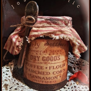 May include: A vintage-style jar with a red and white striped fabric cover and a wooden spoon. The jar is labeled "rly Settlers General Store DRY GOODS" with listed items. The jar sits on a white lace doily.