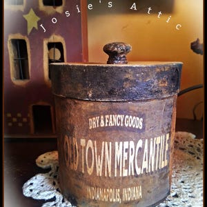 May include: A cylindrical wooden container with a dark, distressed finish and a matching lid featuring a small knob. The front displays the text "DRY & FANCY GOODS", "OLD TOWN MERCANTILE", and "INDIANAPOLIS, INDIANA" in a light, weathered font.