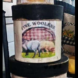 May include: Three round, black-rimmed, white storage containers with red and white gingham patterns. The top container says "Farmhouse Buds", the middle container says "Fine Woolens", and the bottom container says "Fresh Milk". Each container features a different farm animal illustration.