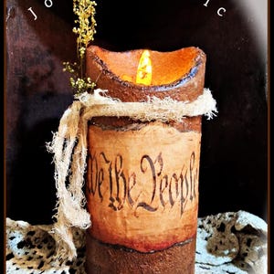May include: A rustic-style candle with a warm, flickering light. The candle features a parchment-like paper wrap with the words "We the People" in elegant script. It is adorned with a fabric wrap and small sprigs of dried flowers.
