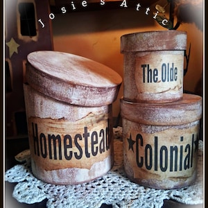 May include: Three round, lidded containers with a distressed, weathered brown and white finish. The largest container is labeled "Homestead," while the two smaller stacked containers are labeled "The Olde" and "Colonial" with a black star graphic.