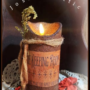 May include: A rustic candle with a flickering flame, wrapped with twine and adorned with dried sprigs. The candle has a textured brown surface and a label that reads "THE KEEPING ROOM". A red flower and lace add to the cozy aesthetic.
