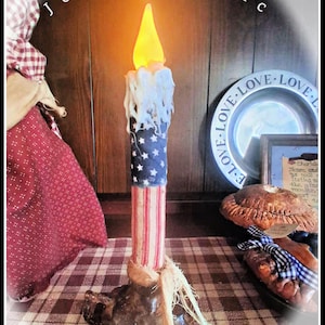 May include: A decorative candle with a patriotic theme. The candle features a blue section with white stars, a red and white striped section, and a faux melted wax effect. The candle sits on a dark brown base and has a flickering yellow flame. The text "Josie's Attic" is at the top.