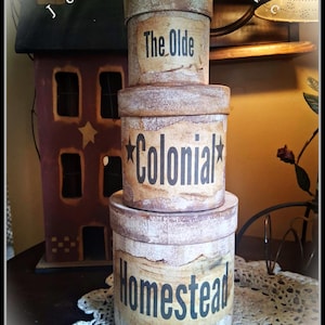 May include: A stack of three cylindrical boxes with a distressed, weathered finish in shades of beige and brown. Each box features black text: the top reads The Olde, the middle reads Colonial with stars, and the bottom reads Homestead.