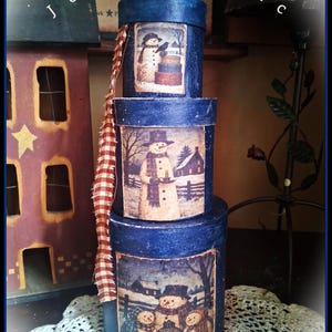 May include: A stack of three cylindrical blue boxes with a textured finish. Each box features a rectangular print of snowmen in winter scenes. A frayed red and white checkered fabric strip hangs from the top box. The text Josies Attic is visible.