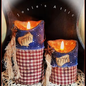May include: Two decorative candles with a patriotic theme. The candles have a red and white checkered base, topped with a blue fabric section with white stars and the number "1776". Each candle has a faux flame and a star accent.