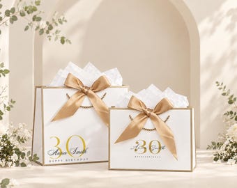 Pode incluir: Duas sacolas de presente retangulares brancas com borda dourada, cada uma com um número 30 grande em dourado, o nome Anna Smith em letra cursiva preta e o texto HAPPY BIRTHDAY. Cada sacola é adornada com um grande laço de cetim dourado e papel de seda branco.