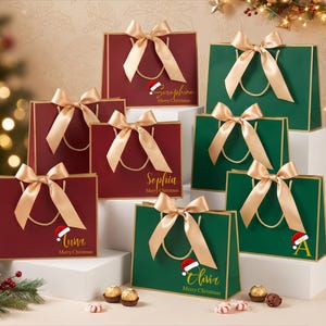 Op de afbeelding: Een collectie rechthoekige cadeauzakjes in donkerrood en bosgroen, elk voorzien van een goudkleurige strik, gouden koordhengsels en gepersonaliseerde namen in gouden schrijfletters met de tekst Merry Christmas en een kleine kerstmuts-afbeelding.