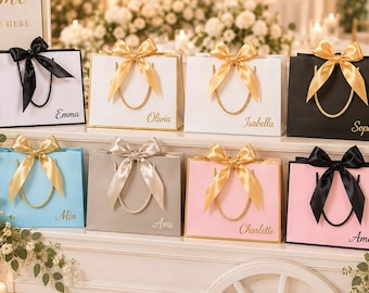 Pode incluir: Uma coleção de oito sacos de presente retangulares em branco, preto, azul claro, cinza e rosa, cada um com um laço de fita de cetim, alças trançadas e um nome personalizado em letra cursiva: Emma, Olivia, Isabella, Sophia, Mia, Ava, Charlette e Amelia.
