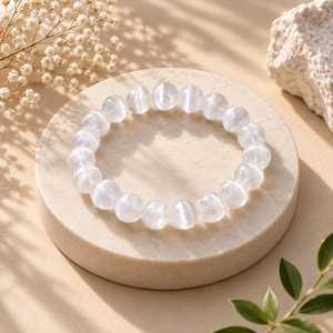 Op de afbeelding: Een elastische armband bestaande uit ronde, doorschijnende witte kralen met een subtiele chatoyante glans. De kralen zijn van gelijke grootte en gerangschikt in een cirkelvormige lus, waarbij ze het licht reflecteren op hun gladde oppervlakken.