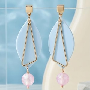 Könnte beinhalten: Ein Paar Ohrhänger mit einem hellblauen, mandelförmigen Element, einem goldfarbenen dreieckigen Rahmen und einer kleinen, runden, durchscheinenden rosa Perle am unteren Ende. Die Ohrringe sind an goldfarbenen Stiften mit Verschluss befestigt.