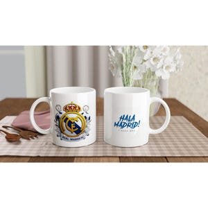 Op de afbeelding: Twee witte keramische mokken met Real Madrid branding. Eén mok toont het gouden, blauwe en witte Real Madrid FC embleem met voetballen. De andere mok bevat de blauwe tekst "HALA MADRID! Y NADA MÁS" in een gestileerd lettertype.