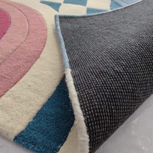 Könnte beinhalten: Ein getufteter Teppich mit einem Muster aus rosa und blauen konzentrischen Halbkreisen sowie einem blau-weißen Schachbrettmuster. Der Teppich ist umgeschlagen und zeigt eine dunkelgraue, strukturierte, rutschfeste Rückseite und eine hellblaue Kante.