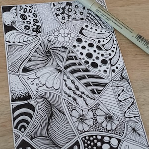Op de afbeelding: Een rechthoekig wit vel papier met een zwarte inkttekening in zentangle-stijl, bestaande uit diverse geometrische patronen, bloemmotieven, stippen en golvende lijnen. Een lichte Sakura Pigma Micron pen met tekst ligt diagonaal op het papier.