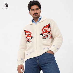 May include: A cream-colored knit zip-up cardigan featuring a shawl collar and two front pockets. The garment displays a red and black fox head graphic on each side of the chest, with small blue paw print patterns near the ribbed cuffs and hem.