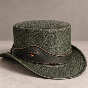 Peut inclure: Un haut-de-forme avec un motif texturé en peau de crocodile vert olive. Une bande en cuir marron foncé entoure la base, agrémentée d'un détail d'œil orange réaliste incrusté dans le cuir, créant un point focal saisissant.