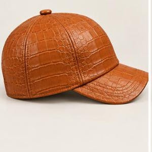 May include: A tan baseball cap featuring a textured crocodile skin pattern across the entire surface. The cap has a rounded crown with a small button at the top and a curved brim extending from the front, constructed from multiple panels.