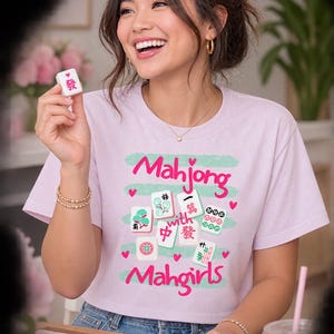 May include: A light purple short-sleeved t-shirt featuring a graphic print with the text Mahjong with Mahgirls, surrounded by illustrations of various mahjong tiles and small pink hearts, set against a light green horizontal brushstroke background.