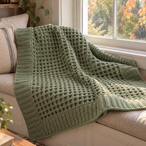 May include: A sage green crochet blanket featuring a textured waffle stitch pattern throughout the center and a wide, ribbed border along the edges. The blanket is draped over a surface, showcasing the intricate yarn work and uniform square grid.