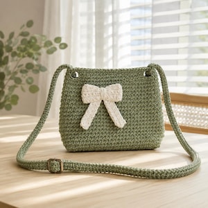 May include: A sage green crocheted crossbody bag featuring a textured stitch pattern and a prominent cream-colored crocheted bow attached to the front center. The bag includes a matching green crocheted shoulder strap with a metal buckle adjustment.