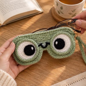 Könnte beinhalten: Ein hellgrünes, gehäkeltes Brillenetui mit zwei großen, kreisförmigen weißen und schwarzen Augen und einem kleinen aufgestickten Lächeln. Das Etui verfügt über eine passende grüne Handschlaufe an der Seite und ist für eine Brille mit dunklem Rahmen.