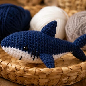 May include: A small, handmade crochet shark toy featuring a dark blue upper body and a white underbelly. The shark has a prominent dorsal fin, side fins, a forked tail, and small black plastic eyes. It is crafted with visible yarn stitches.