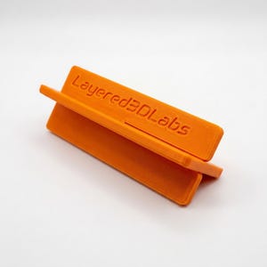 May include: A bright orange, V-shaped stand featuring the embossed text Layered3DLabs on the front panel. The object has a textured surface finish consistent with 3D printing, with two angled rectangular panels joined at the base to form a support.