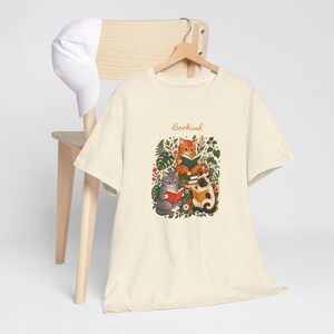 Puede incluir: Una camiseta de manga corta de color crema con un gráfico de tres gatos leyendo libros rodeados de follaje y flores. La palabra Bookish está impresa en letra cursiva naranja sobre los gatos. Una gorra de béisbol blanca cuelga de la silla.