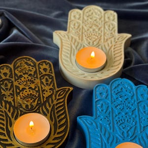 May include: Three Hamsa hand-shaped candle holders in cream, black with gold detailing, and bright blue. Each holder features an intricate embossed floral and geometric pattern and contains a single lit white tea light candle.