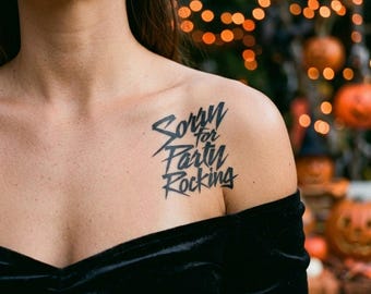 May include: A black temporary tattoo on skin featuring the text Sorry for Party Rocking in a stylized, angular font. The tattoo is positioned on the upper chest area, just below the collarbone, worn with a black off-the-shoulder top.
