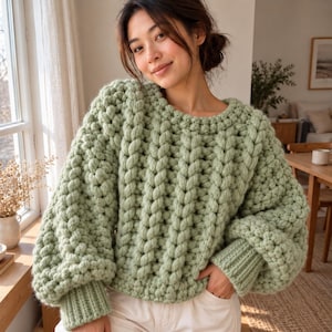 May include: A sage green chunky knit sweater featuring a prominent vertical cable knit pattern, a round neckline, and long sleeves with wide ribbed cuffs. The garment is crafted from thick yarn, creating a textured, voluminous appearance.