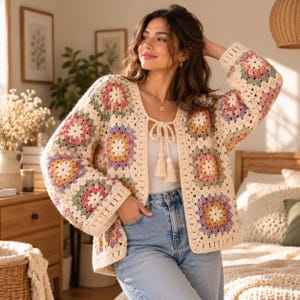 May include: A long-sleeved crochet cardigan made of cream-coloured yarn featuring a pattern of multi-coloured granny squares in shades of pink, yellow, green, and purple. The garment has a front tie closure with two cream-coloured tassels.